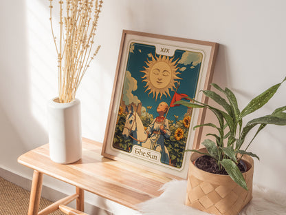 A framed Sun Tarot Card poster rests on a wooden bench, surrounded by a white vase with dried flowers and a potted green plant, creating a cozy, sunlit setting.