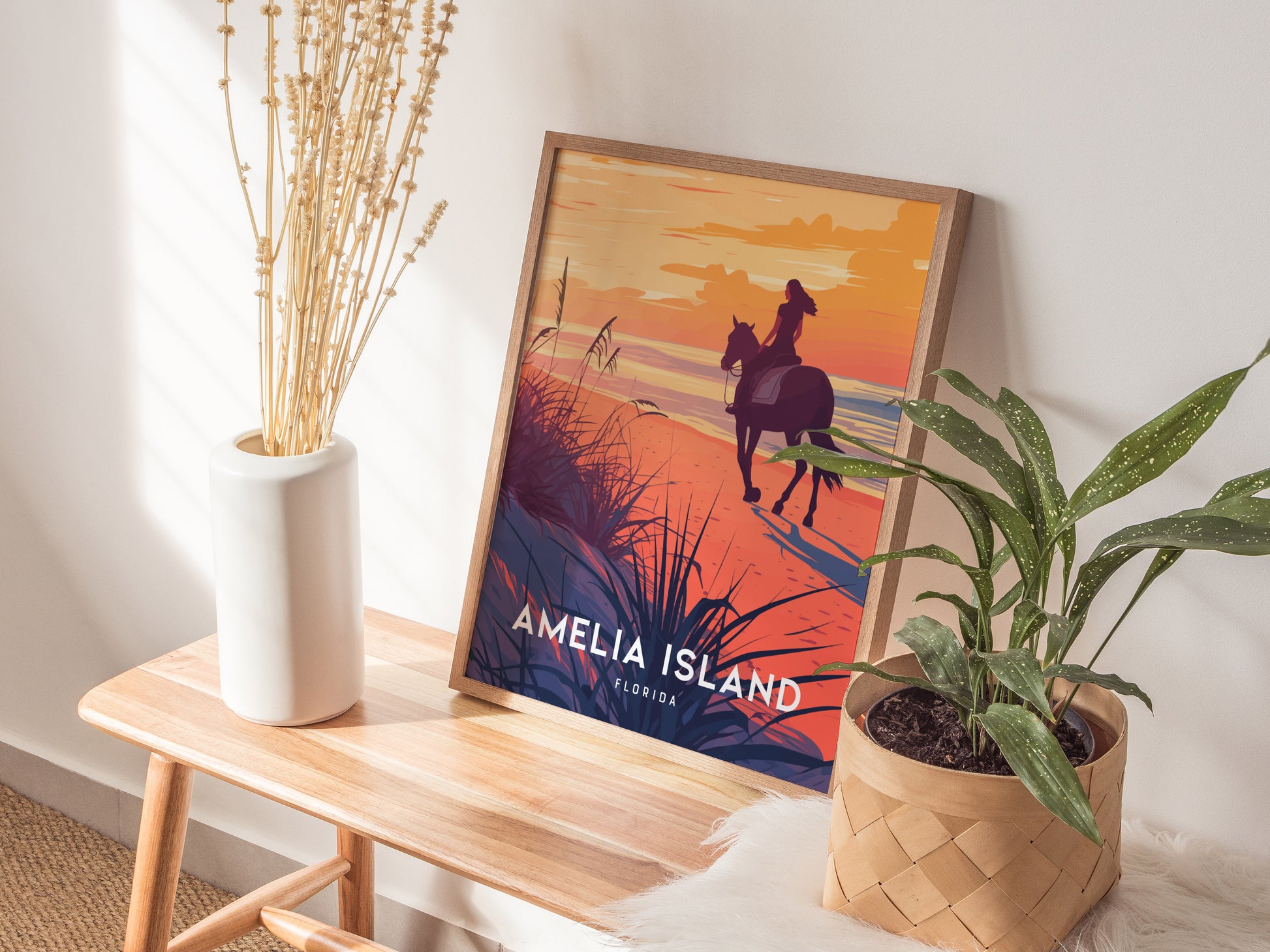 A framed Amelia Island poster featuring a woman on horseback at sunset is placed on a wooden bench, surrounded by a potted plant and a vase with dried flowers.