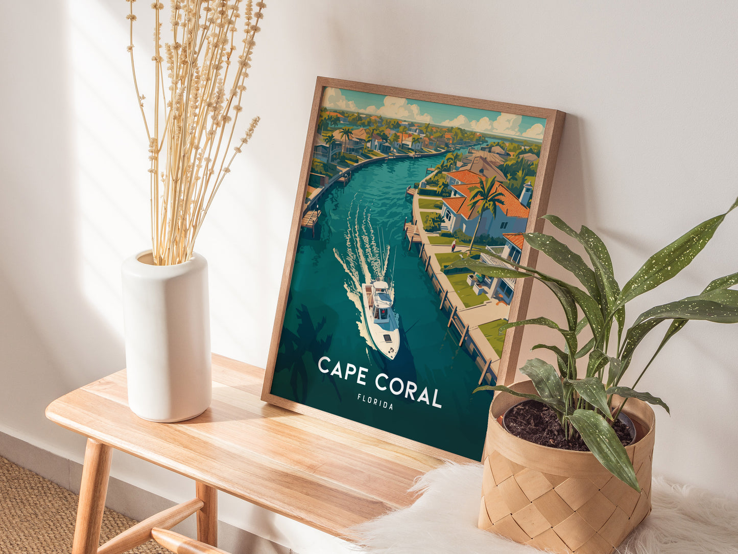 A framed Cape Coral, Florida poster depicting a canal with a boat, tropical homes, and palm trees sits on a wooden bench next to a white vase and a potted.