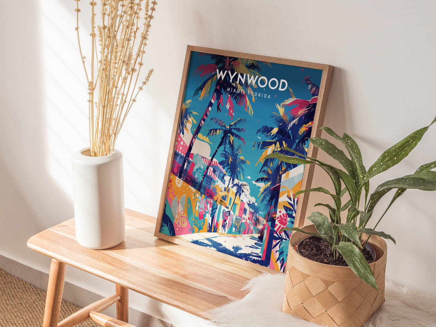 A framed Wynwood Walls Miami poster featuring vibrant street art and palm trees is displayed on a wooden table. The setup includes a white vase with dried flowers and a potted green plant, creating a stylish.