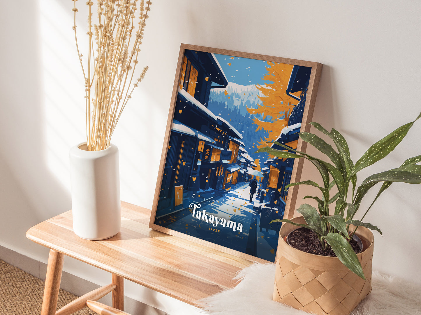 A framed Takayama Japan poster rests on a wooden bench, surrounded by a white vase with dried flowers and a potted green plant, against a softly lit wall.