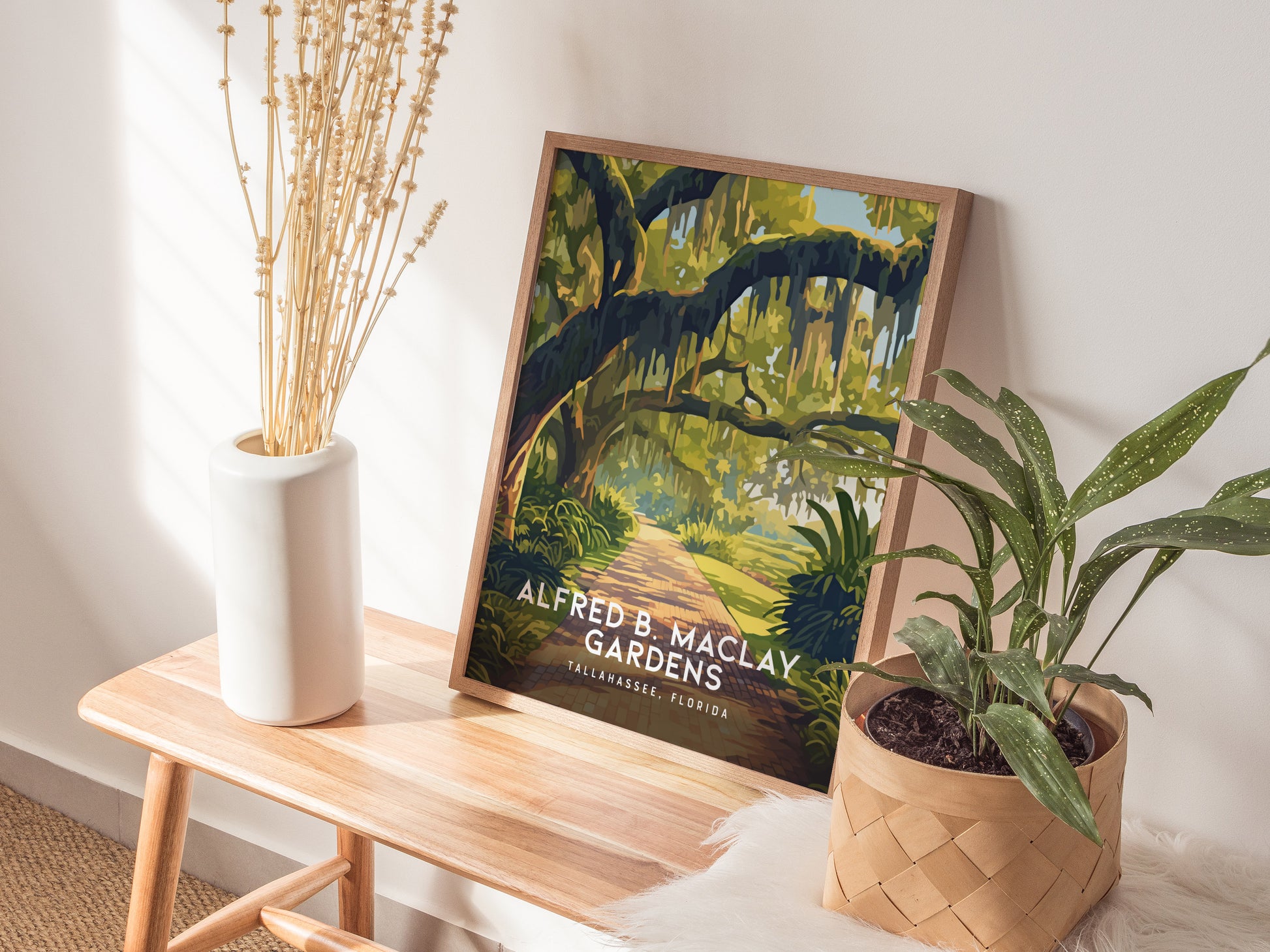 A framed poster of Alfred B. Maclay Gardens is displayed on a wooden bench, accompanied by a white vase with dried flowers and a potted plant in a basket.