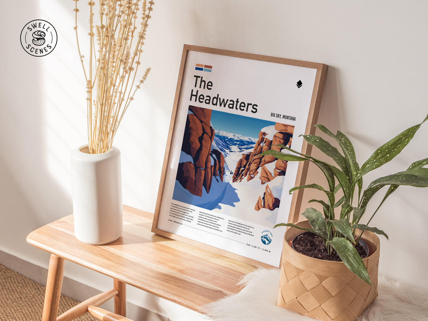 A framed poster titled The Headwaters leans against a wall on a wooden bench, surrounded by a vase with dried stems and a potted plant.