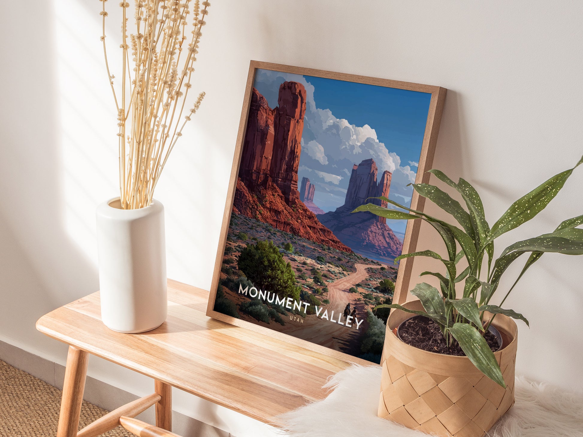 A framed Monument Valley poster featuring red sandstone mesas and a blue sky rests on a wooden bench, accompanied by a white vase with dried stems and a potted plant.