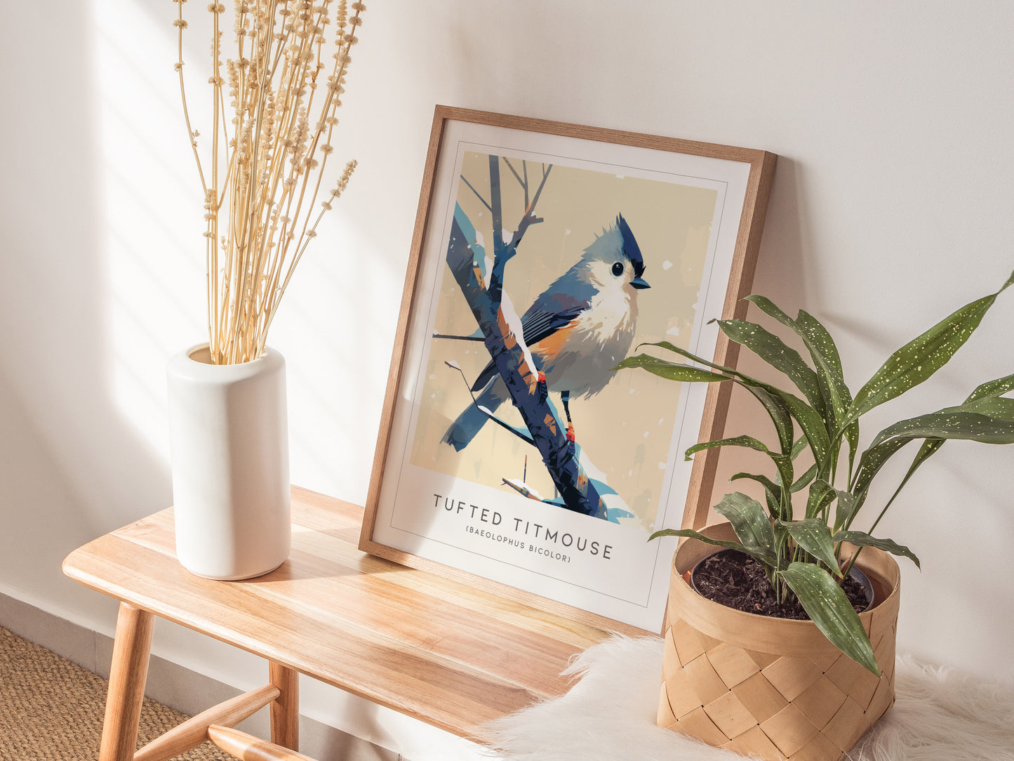 A framed Tufted Titmouse art print rests on a wooden bench, surrounded by a white vase with dried stems and a potted green plant. Sunlight casts soft shadows, creating a cozy, natural ambiance.