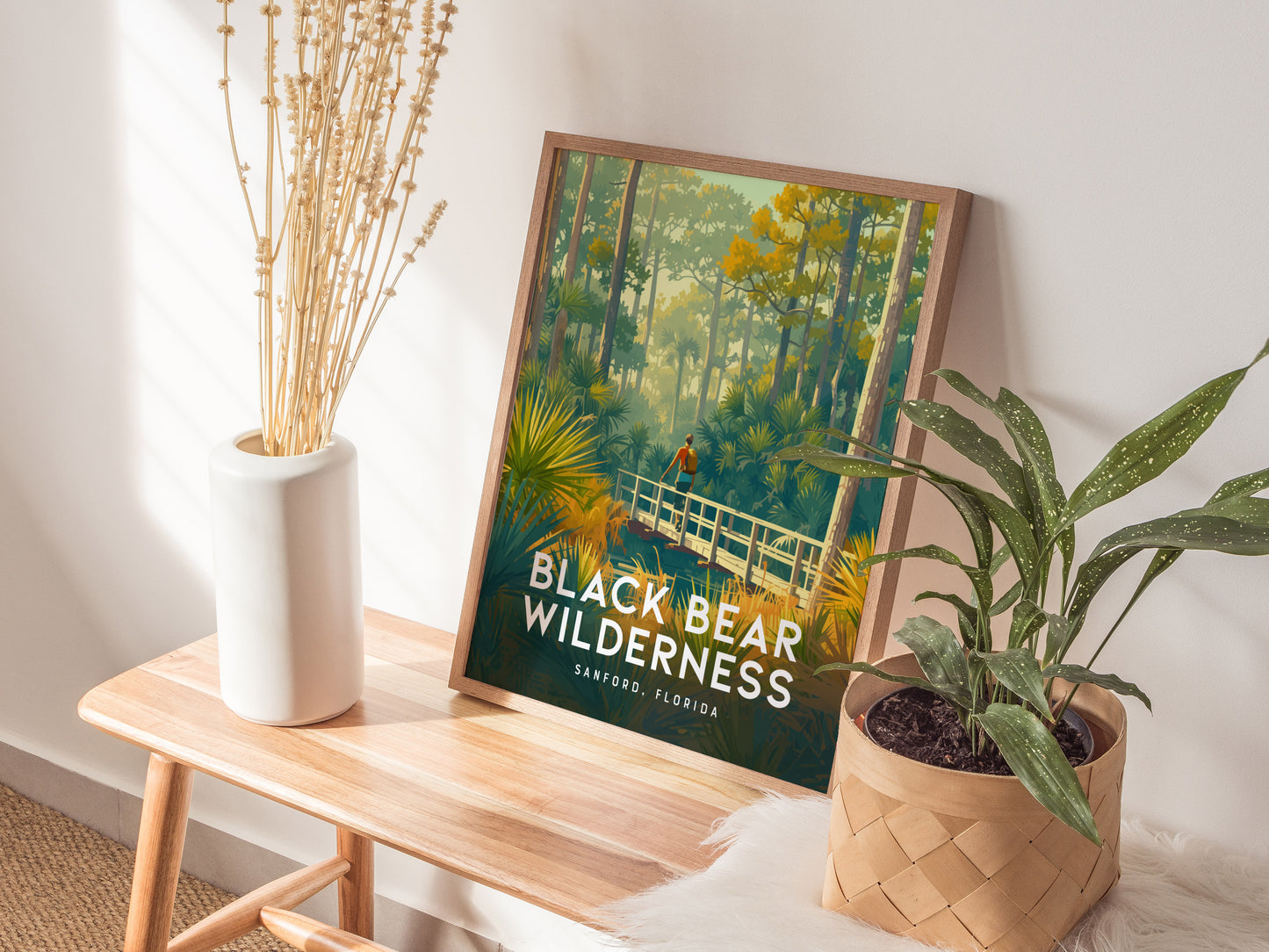 A framed Black Bear Wilderness poster rests on a wooden table, accompanied by a white vase with dried flowers and a potted plant.