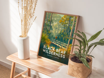 A framed Black Bear Wilderness poster rests on a wooden table, accompanied by a white vase with dried flowers and a potted plant.
