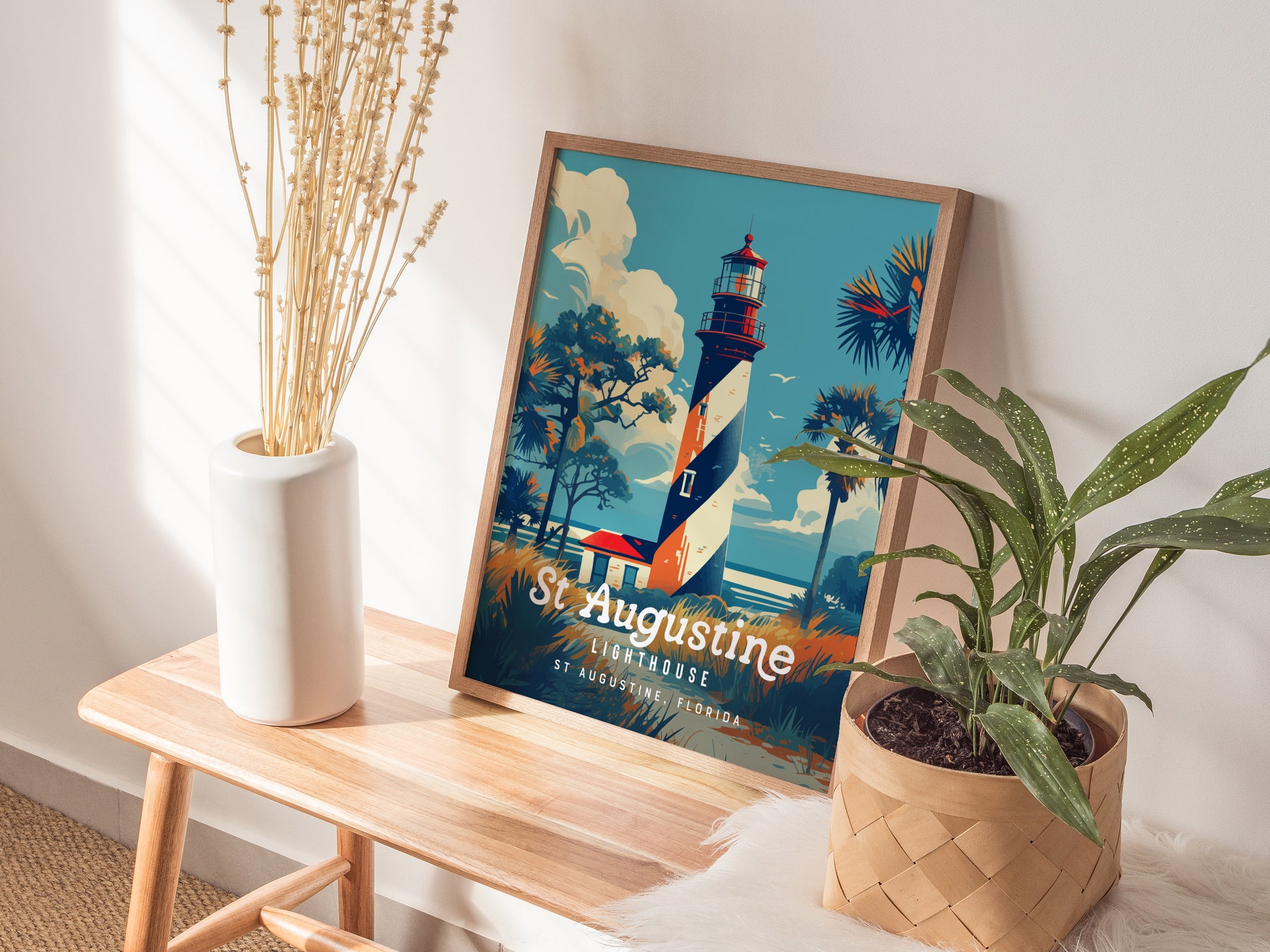 A colorful poster featuring the St. Augustine Lighthouse with a vibrant sky and palm trees is displayed on a wooden table. Nearby, a white vase with dried flowers and a potted plant add decorative touches.