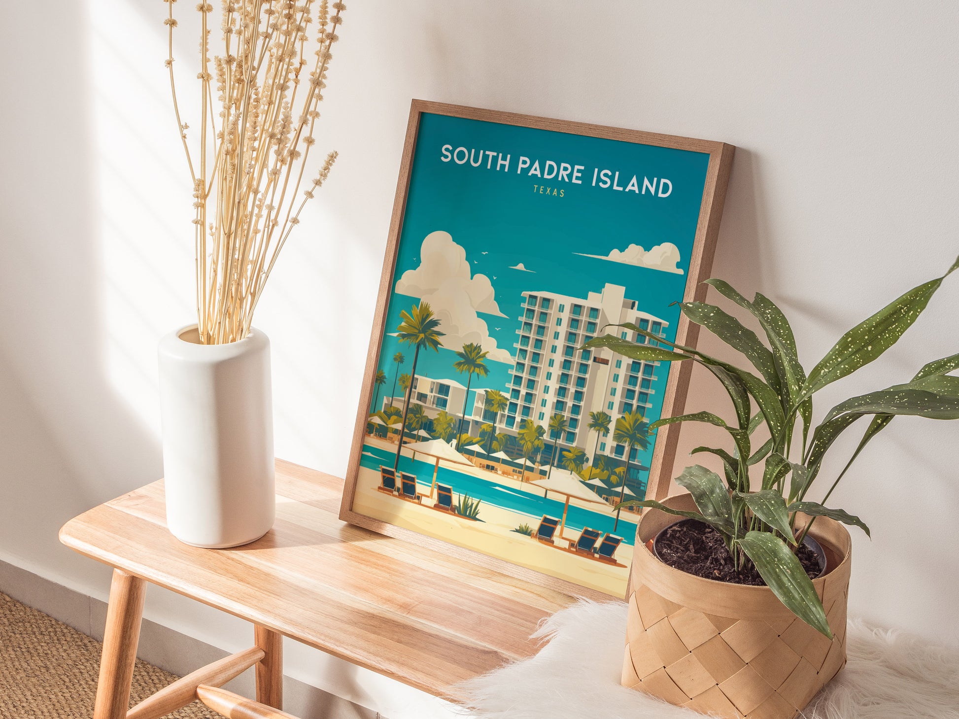 A framed South Padre Island poster leans against a wall on a wooden bench, accompanied by a white vase with dried flowers and a potted green plant.