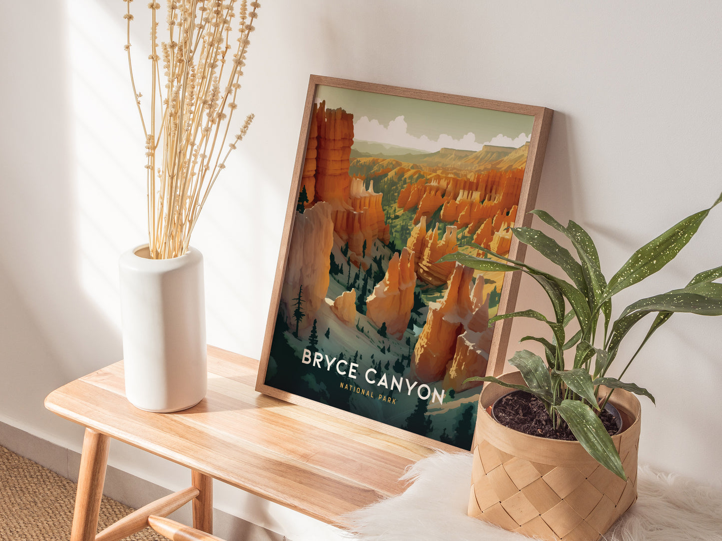 A framed Bryce Canyon National Park print rests on a wooden bench beside a white vase with dried flowers and a potted green plant, all bathed in natural sunlight.