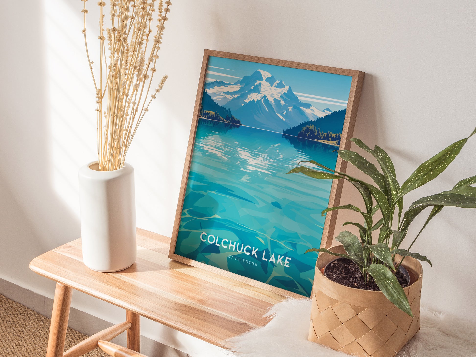 A framed Colchuck Lake poster leans against a wall on a wooden bench, flanked by a white vase with dried stems and a potted green plant.