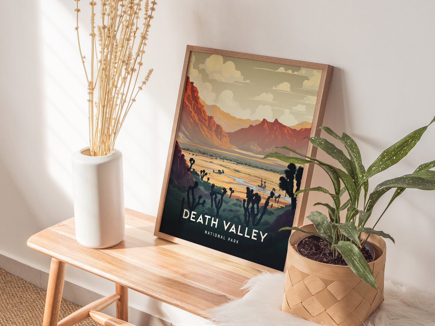A framed Death Valley National Park print is displayed on a wooden bench, accompanied by a white vase with dried stems and a potted plant. Sunlight filters through casting shadows.