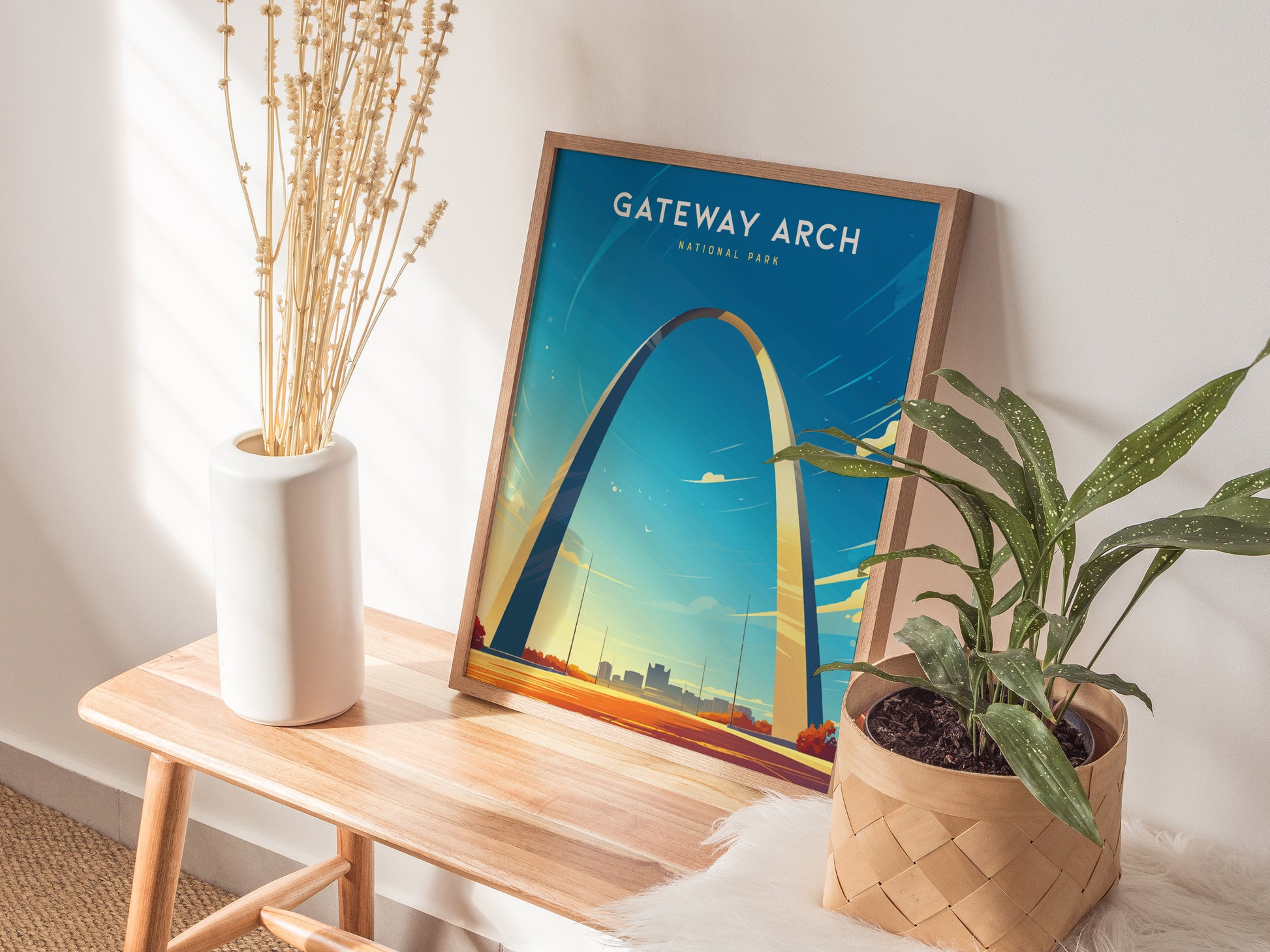 A framed Gateway Arch poster rests on a wooden bench, surrounded by a white vase with dried flowers and a potted green plant, creating a cozy, natural setting.