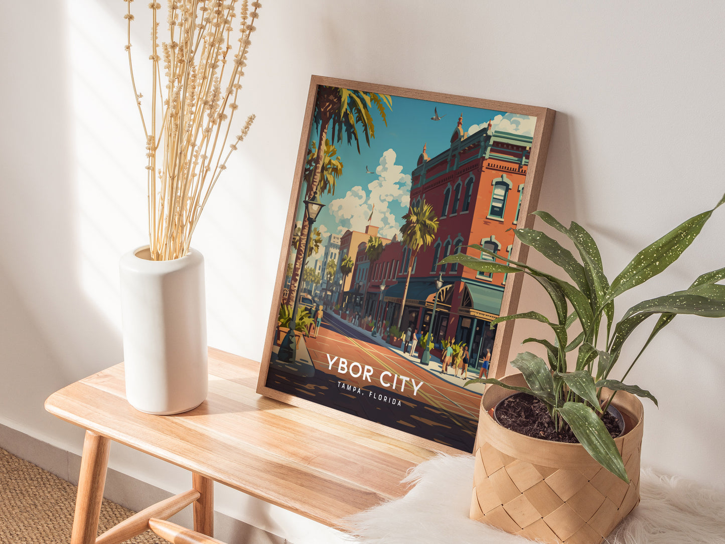 A framed Ybor City, Tampa, Florida poster rests on a wooden bench beside a white vase with dried flowers and a potted plant. Sunlight casts shadows on the wall.