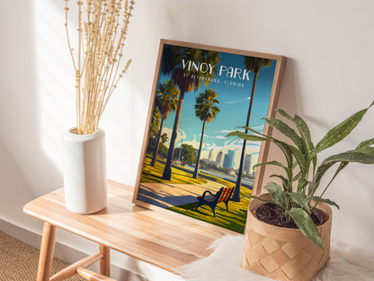 A framed Vinoy Park poster leans against a wall on a wooden bench, flanked by a vase of dried flowers and a potted plant.