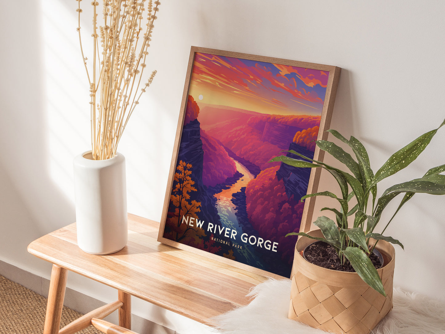 A framed New River Gorge poster with vibrant sunset colors is displayed on a wooden bench, accompanied by a potted plant and a white vase with dried stems.