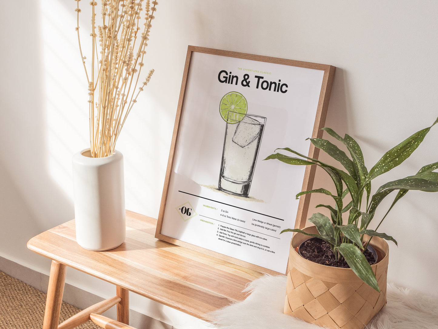 A framed Gin and Tonic Cocktail Poster rests on a wooden bench beside a white vase with dried flowers and a potted green plant, in a sunlit room.