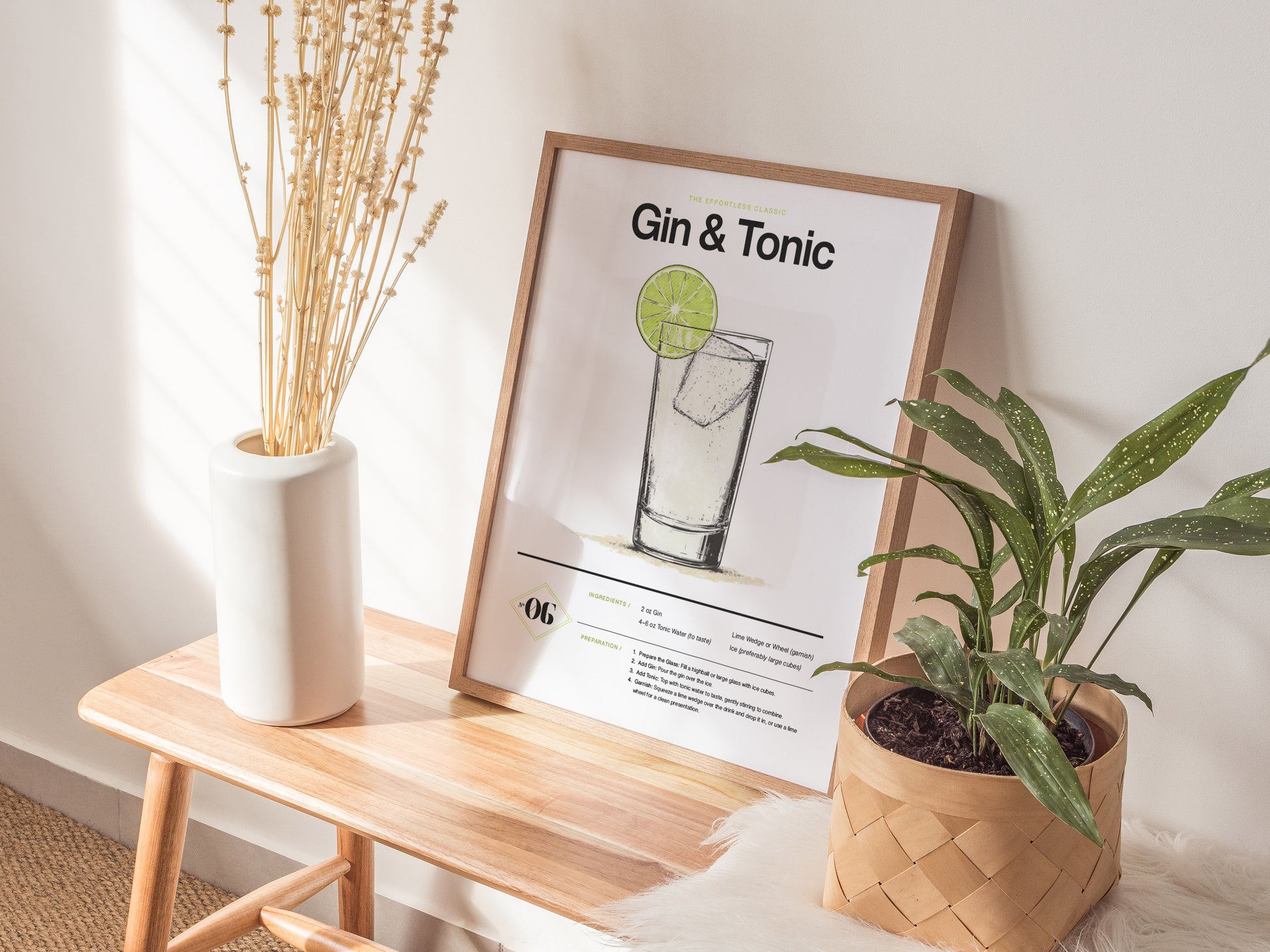 A framed Gin and Tonic Cocktail Poster rests on a wooden bench beside a white vase with dried flowers and a potted green plant, in a sunlit room.
