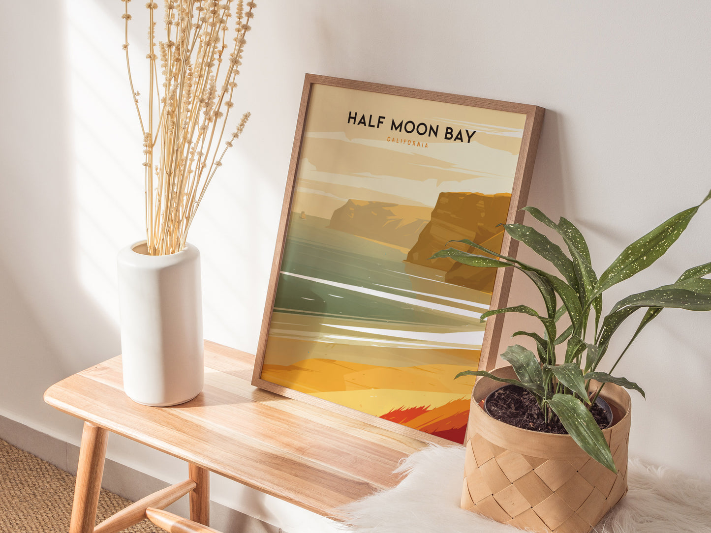 A framed Half Moon Bay poster leans against a wall on a wooden bench, accompanied by a white vase with dried flowers and a potted plant.