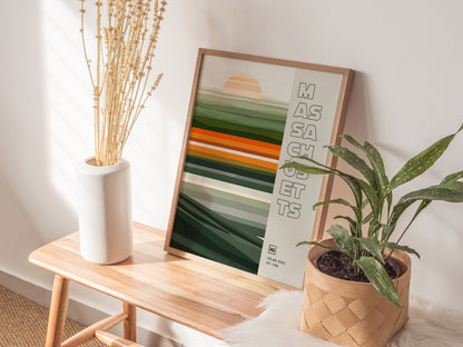 A framed Massachusetts poster with abstract green and orange stripes leans against a wall. Its placed on a wooden bench next to a white vase with dried flowers and a.