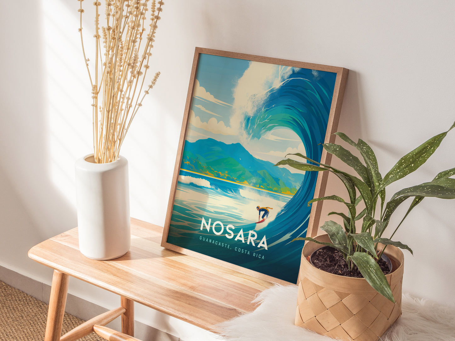 A framed Nosara Costa Rica poster featuring a surfer riding a large wave is placed on a wooden bench next to a white vase with dried flowers and a potted.