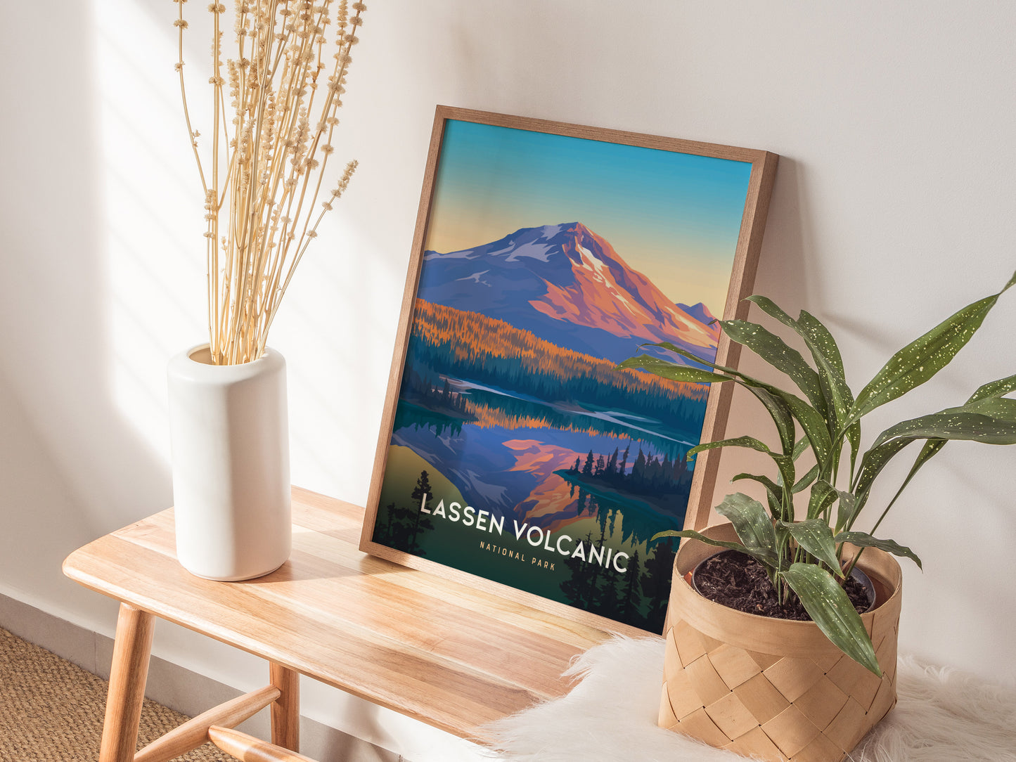 A framed Lassen Volcanic National Park poster leans against a wall, featuring a colorful mountain scene. Its placed on a wooden bench with a potted plant and a white vase.