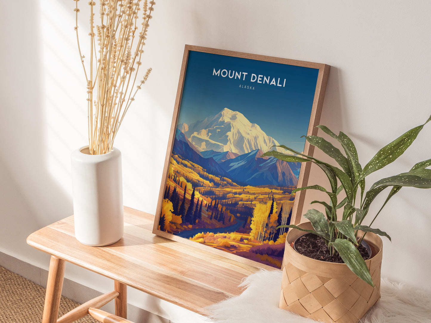 A Mount Denali poster in a wooden frame stands on a wooden bench, accompanied by a white vase with dried flowers and a potted plant in a woven basket.