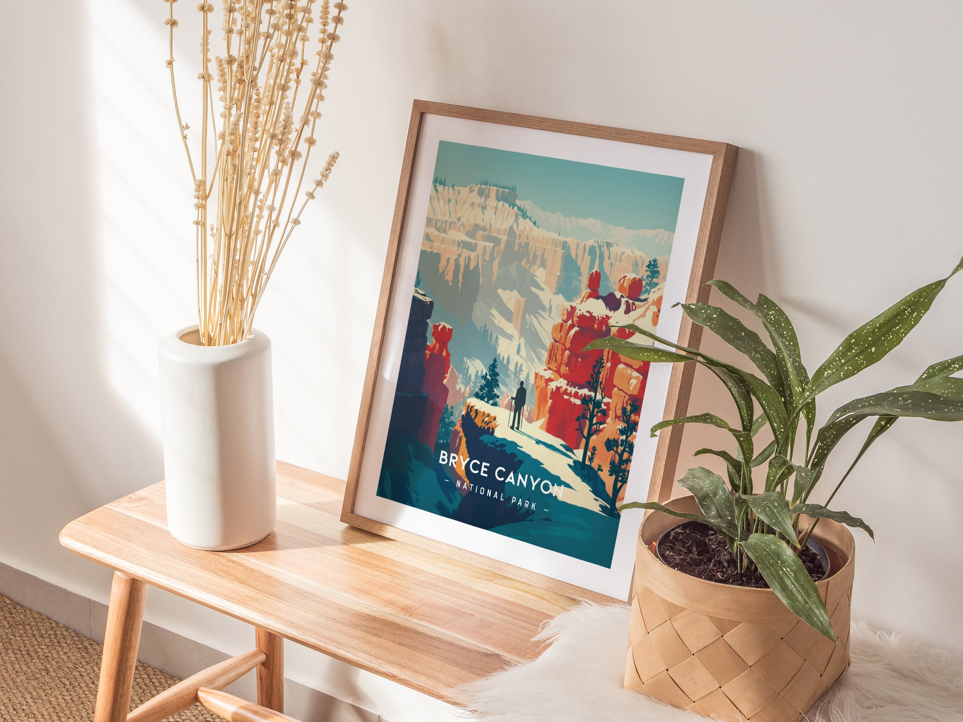 A Bryce Canyon National Park print in a wooden frame rests on a wooden bench, surrounded by a white vase with dried plants and a green potted plant.