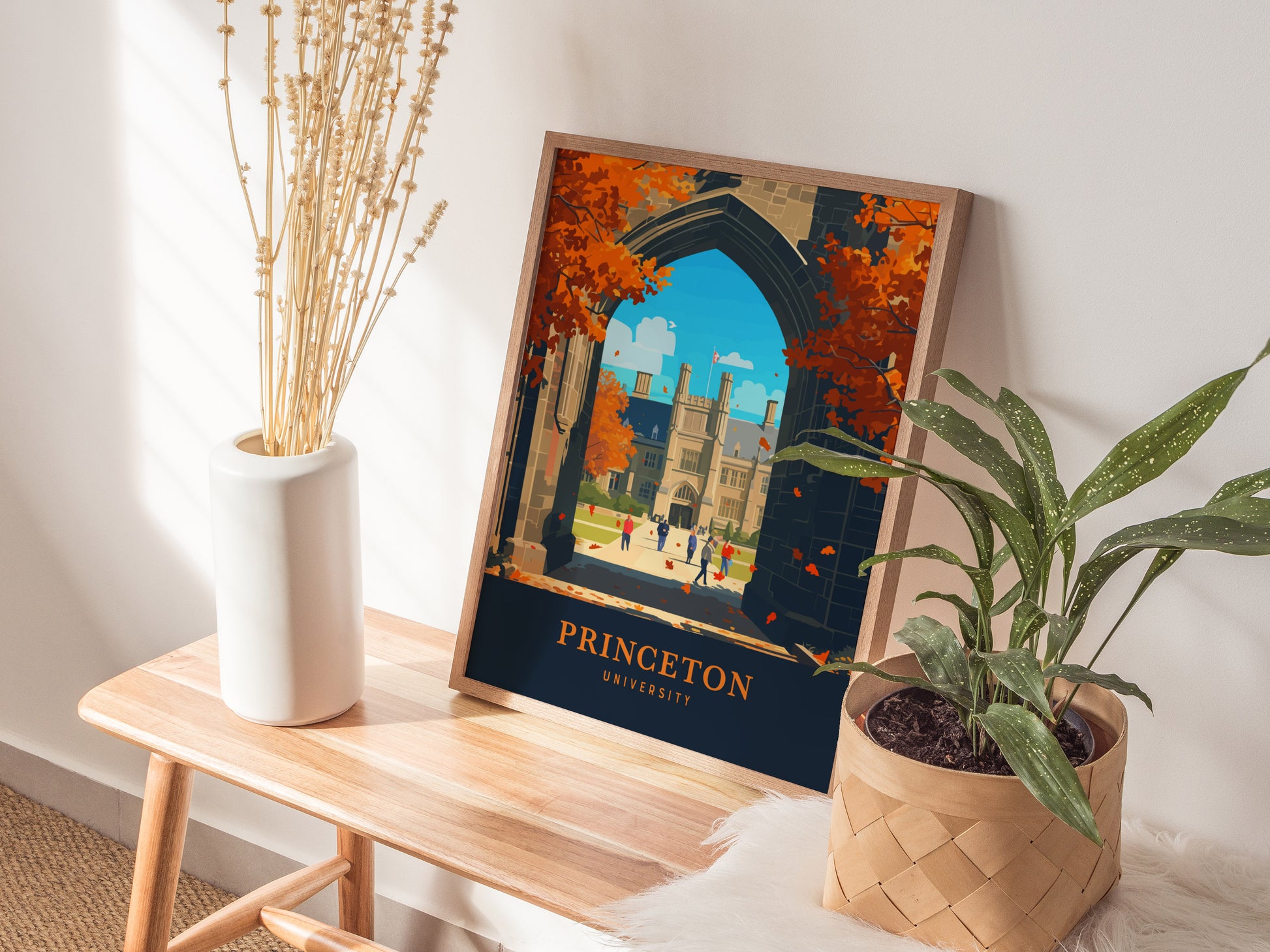 A framed Princeton University poster featuring Gothic architecture and autumn foliage is displayed on a wooden table, accompanied by a white vase with dried flowers and a potted plant.