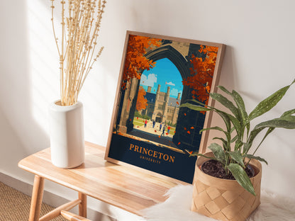 A framed Princeton University poster featuring Gothic architecture and autumn foliage is displayed on a wooden table, accompanied by a white vase with dried flowers and a potted plant.