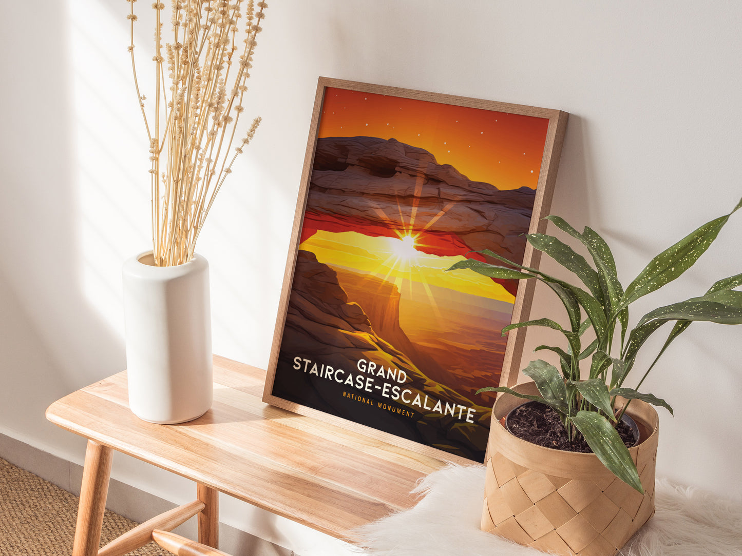 A framed Grand Staircase–Escalante poster featuring a sunset arch is displayed on a wooden bench beside a white vase with dried flowers and a green potted plant.