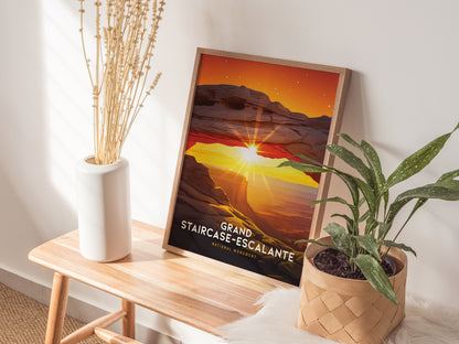 A framed Grand Staircase–Escalante poster featuring a sunset arch is displayed on a wooden bench beside a white vase with dried flowers and a green potted plant.