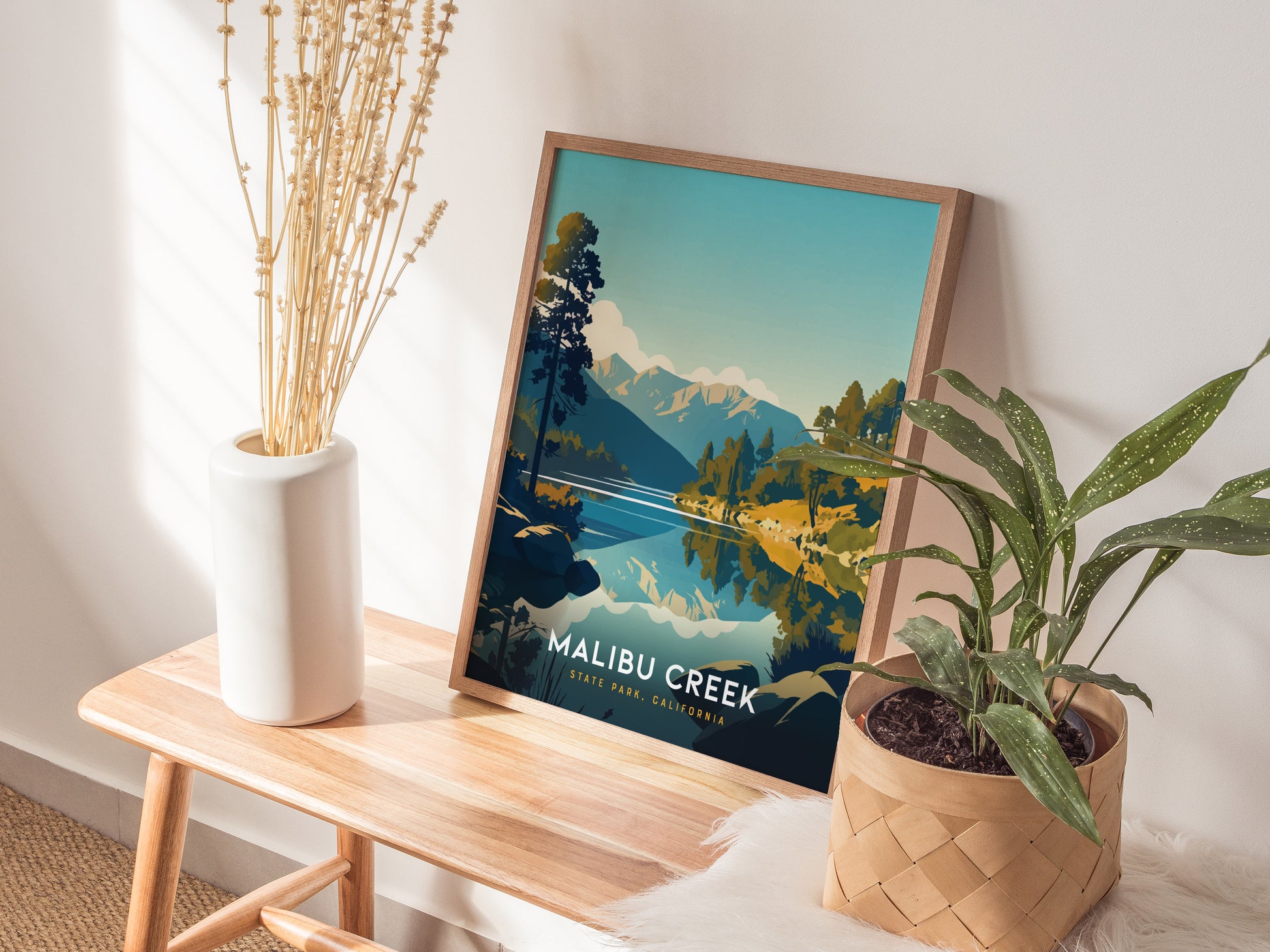 A framed poster of Malibu Creek State Park leans against a wall, accompanied by a white vase with dried flowers and a potted plant on a wooden bench.