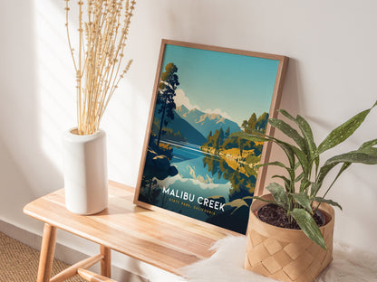 A framed poster of Malibu Creek State Park leans against a wall, accompanied by a white vase with dried flowers and a potted plant on a wooden bench.