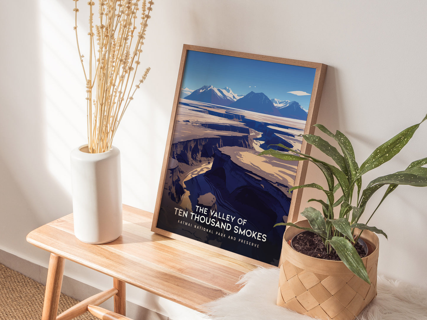 A framed Valley of Ten Thousand Smokes poster leans against a wall on a wooden bench, accompanied by a white vase with dried flowers and a potted plant.