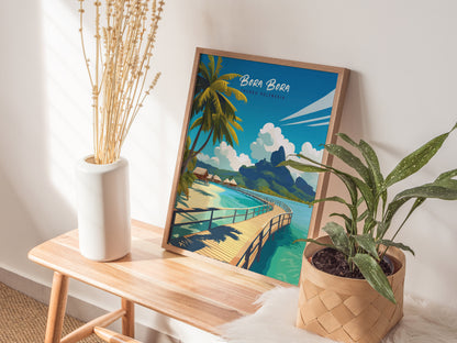 A framed Bora Bora travel print rests on a wooden table, accompanied by a white vase with dried stems and a potted green plant, creating a tropical decor scene.