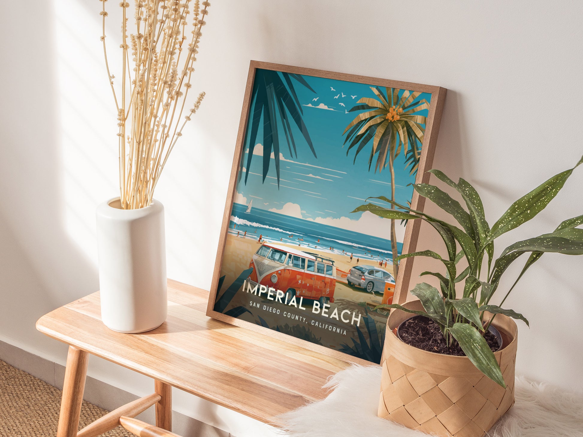 A framed Imperial Beach poster sits on a wooden bench beside a white vase with dried flowers and a potted plant, capturing a sunny coastal scene.