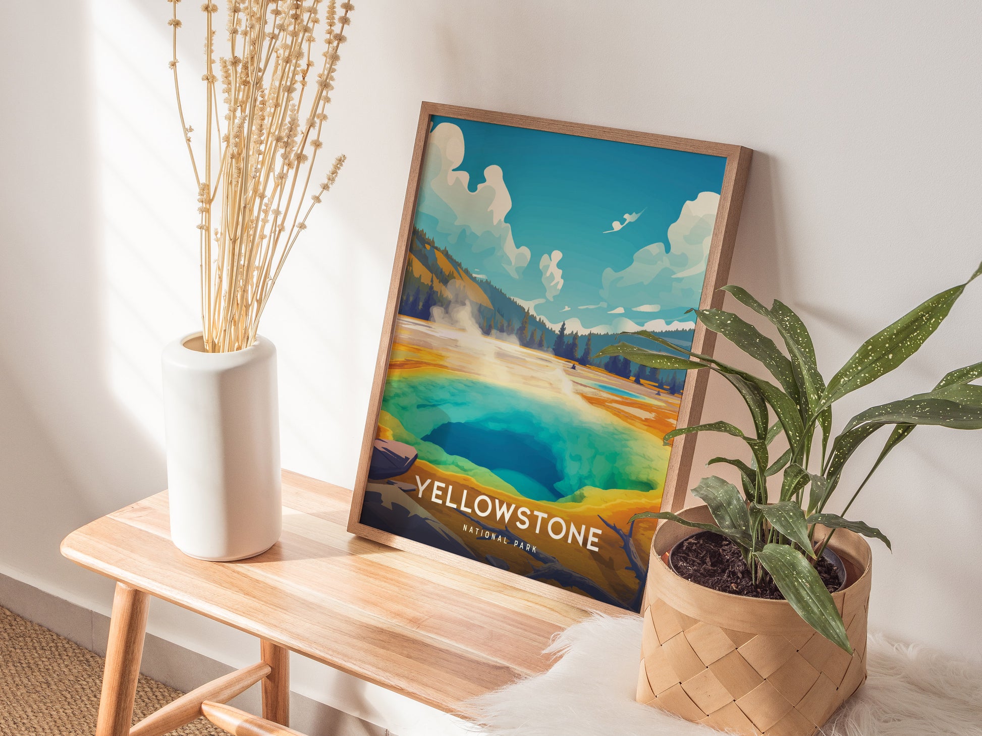 A framed Yellowstone National Park poster featuring the vibrant Morning Glory Pool rests on a wooden bench, accompanied by a white vase with dried flowers and a potted plant.