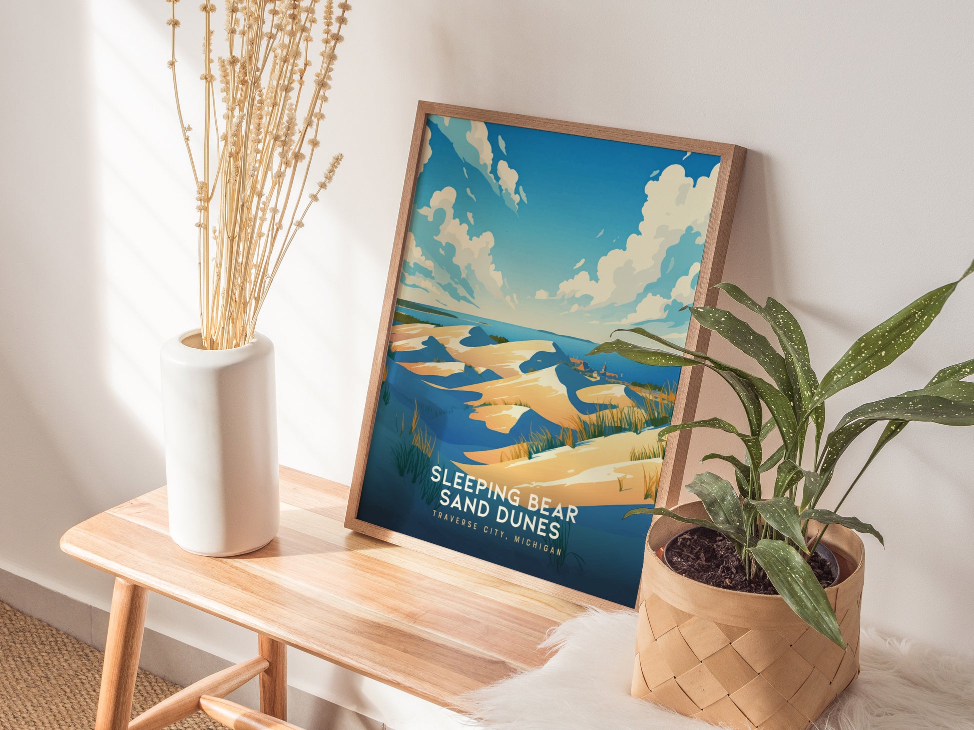 A framed Sleeping Bear Sand Dunes poster leans on a wall, surrounded by a white vase with dried flowers and a potted green plant on a wooden bench.