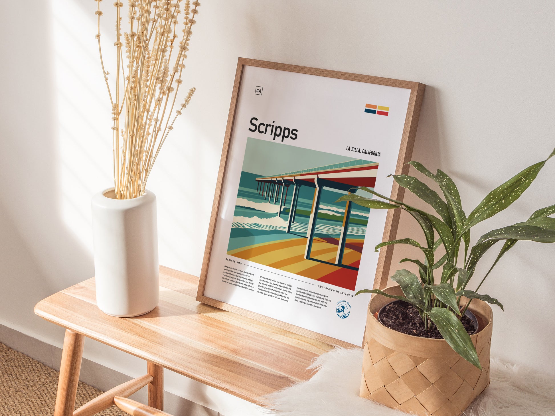 A framed poster of Scripps Pier is displayed on a wooden table, accompanied by a white vase with dried flowers and a potted green plant.