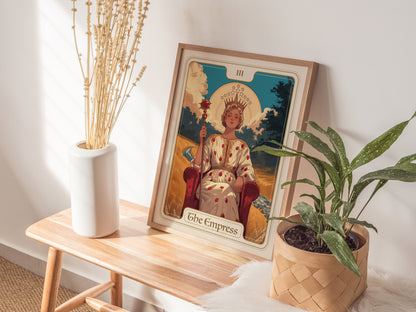 A framed Empress tarot card poster rests on a wooden table, flanked by a white vase with dried flowers and a potted green plant, creating a serene atmosphere.