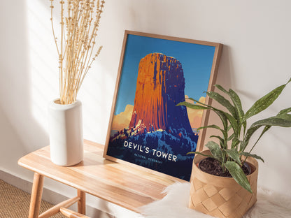 A framed Devils Tower poster leans on a wooden bench beside a white vase with dried flowers and a potted green plant, creating a cozy corner display.