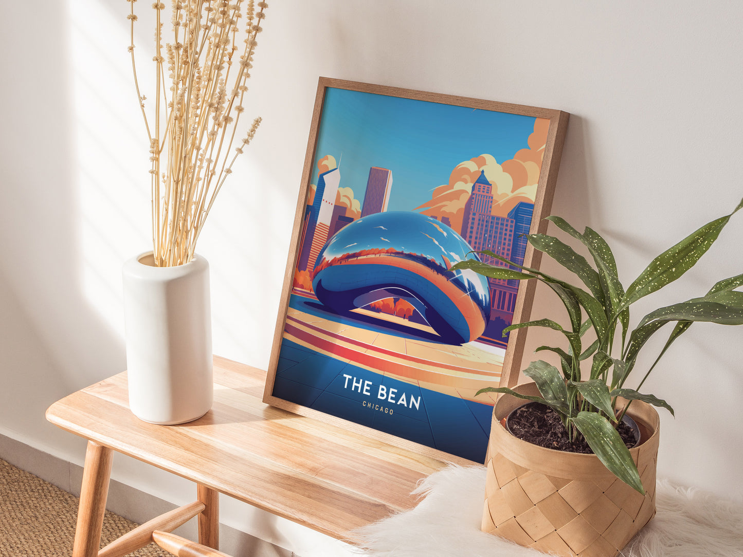 A framed poster of Chicagos Cloud Gate sculpture rests on a light wood table, flanked by a white vase with dried stems and a potted green plant.