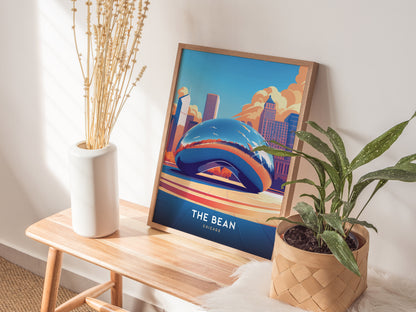 A framed poster of Chicagos Cloud Gate sculpture rests on a light wood table, flanked by a white vase with dried stems and a potted green plant.