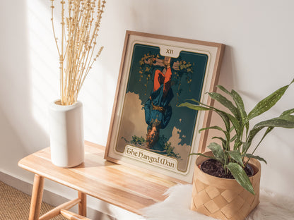A framed Hanged Man tarot card poster is propped on a wooden table beside a white vase with dried flowers and a potted green plant.