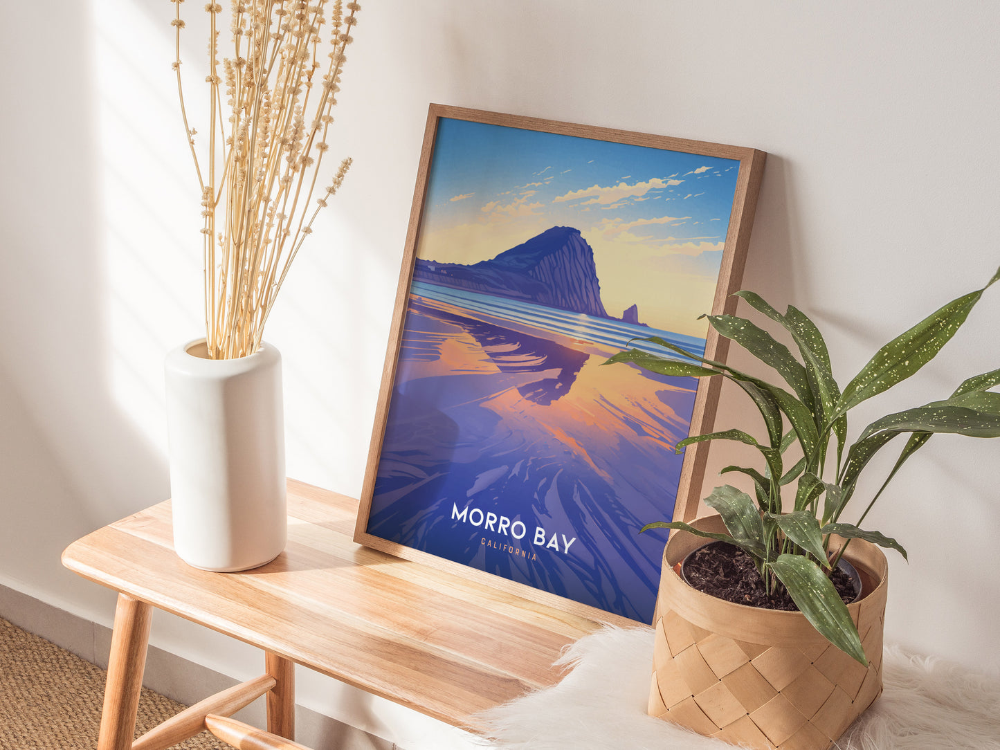 A framed Morro Bay poster with a vibrant coastal scene is displayed on a wooden bench, accompanied by a white vase with dried flowers and a potted plant.