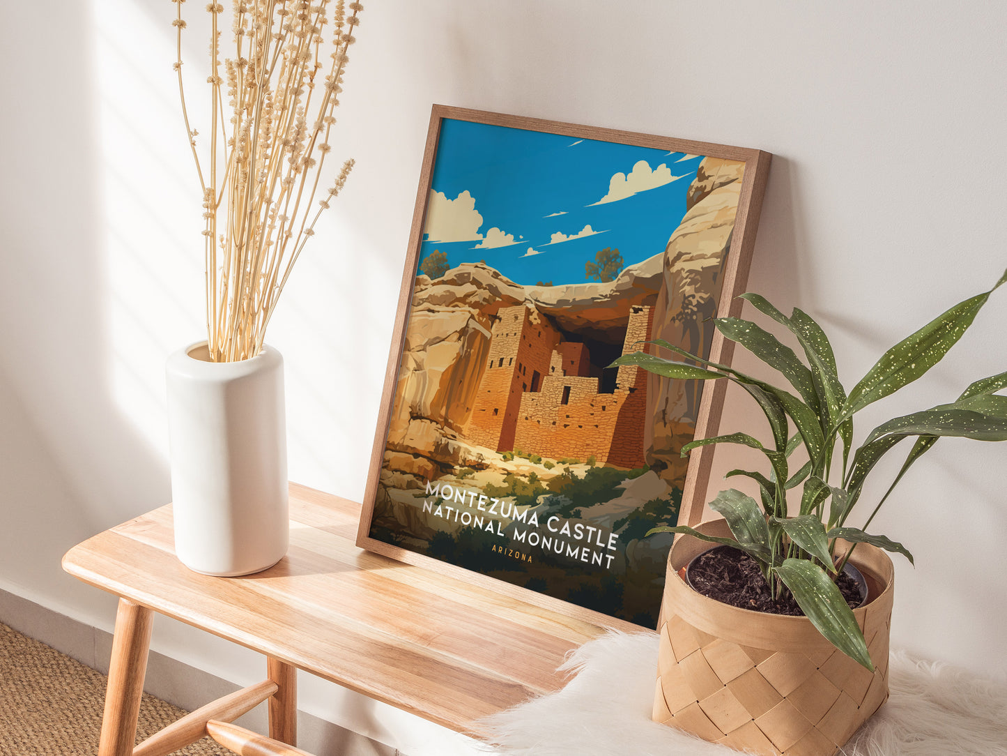 A framed Montezuma Castle National Monument poster rests on a wooden bench next to a white vase with dried flowers and a potted green plant, creating a cozy interior setting.