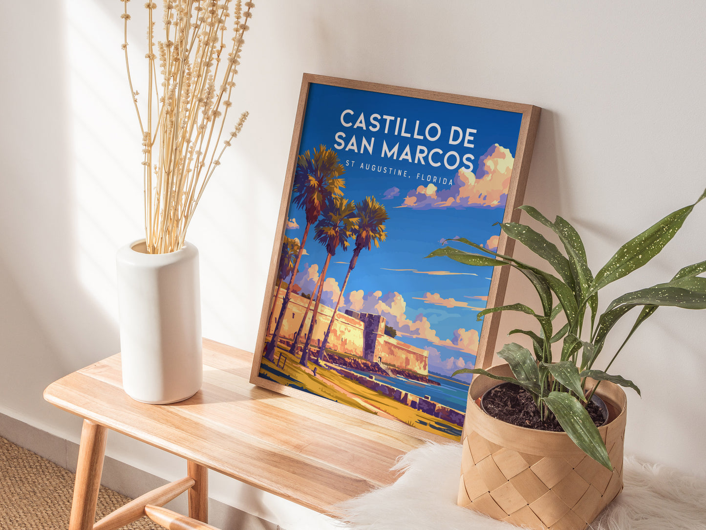 A framed Castillo de San Marcos poster leans on a wall, placed on a wooden bench beside a white vase with dried flowers and a potted plant.