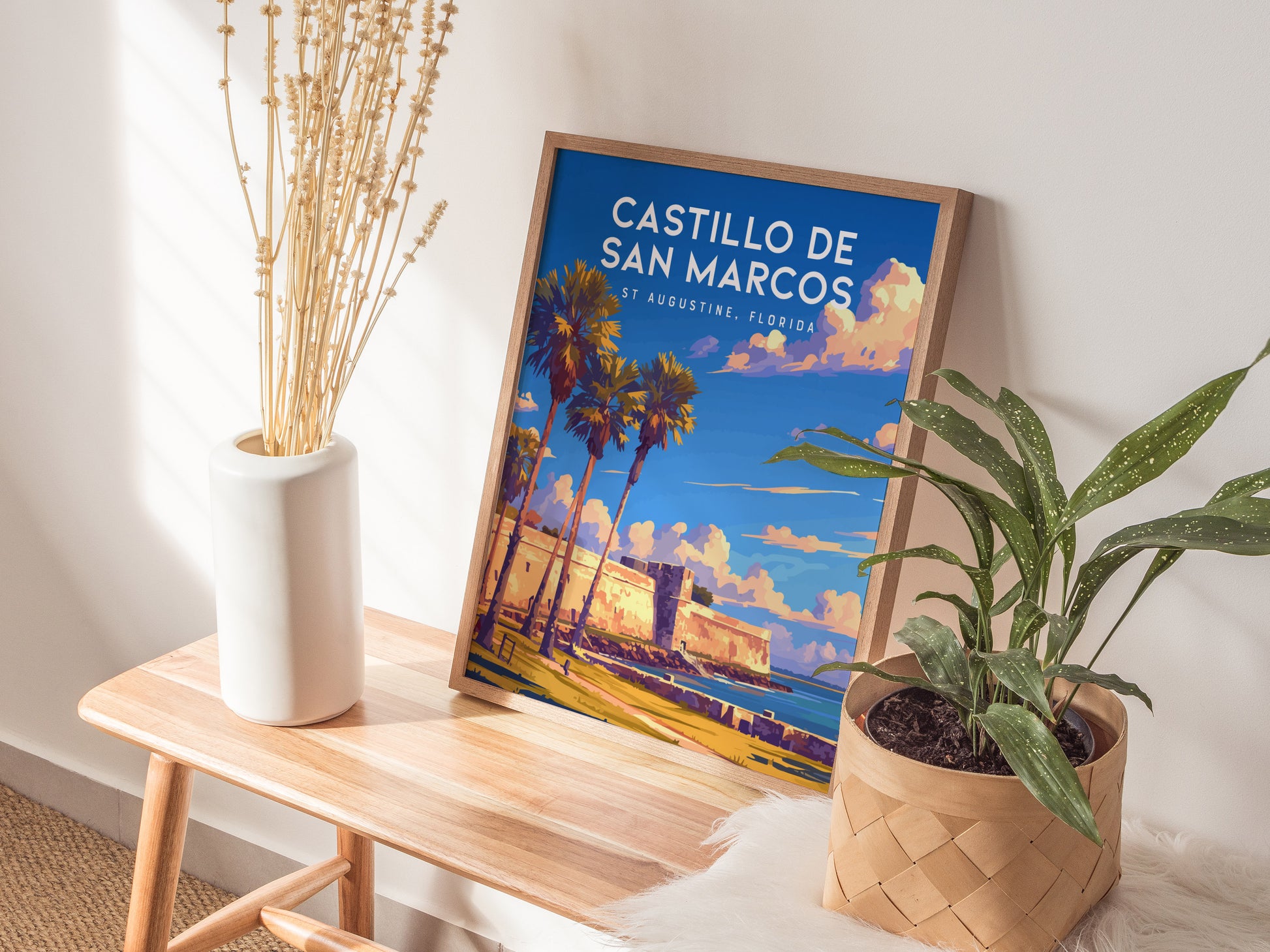 A framed Castillo de San Marcos poster leans on a wall, placed on a wooden bench beside a white vase with dried flowers and a potted plant.