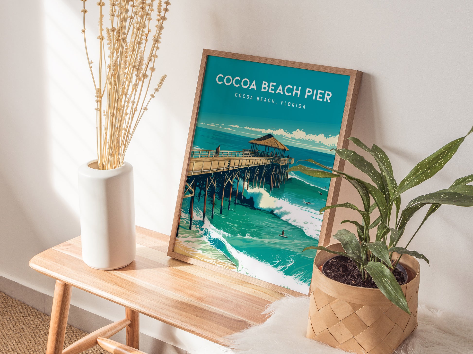 A framed Cocoa Beach Pier poster rests on a wooden table beside a white vase with dried flowers and a potted plant, illuminated by sunlight.